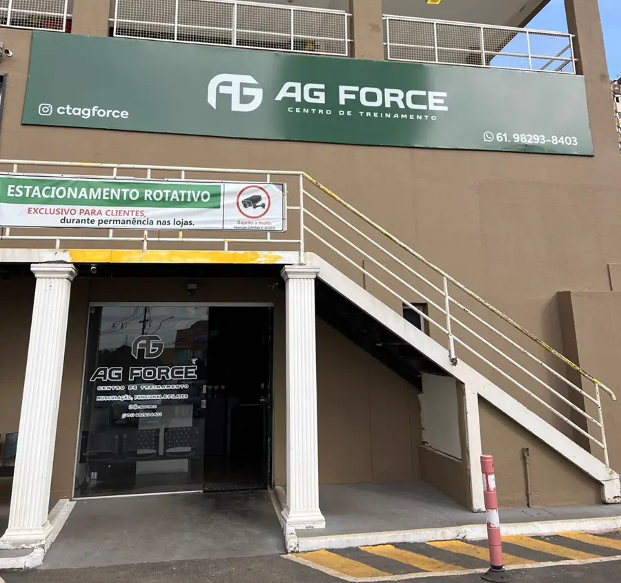 Entrada da AG Force: placa verde com o nome, escada externa branca e porta de vidro embaixo da escada, ao lado da placa do estacionamento rotativo